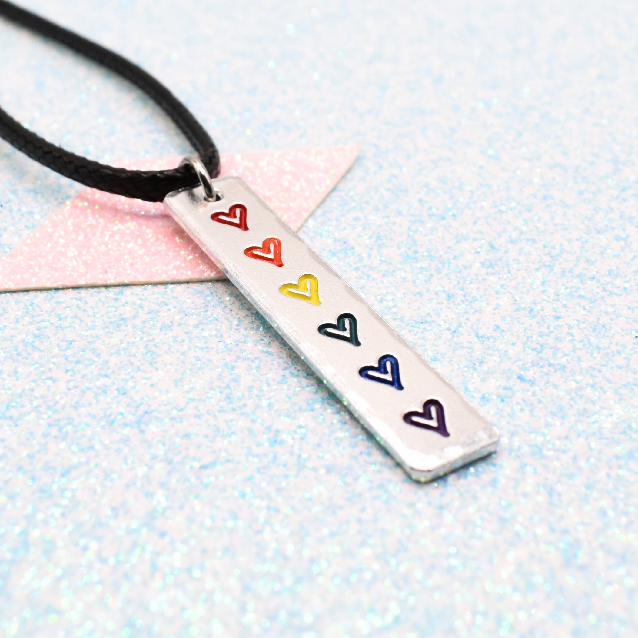 Necklace with a bar pendant on a faux leather cord featuring colourful hearts representing the traditional pride flag on a light blue background