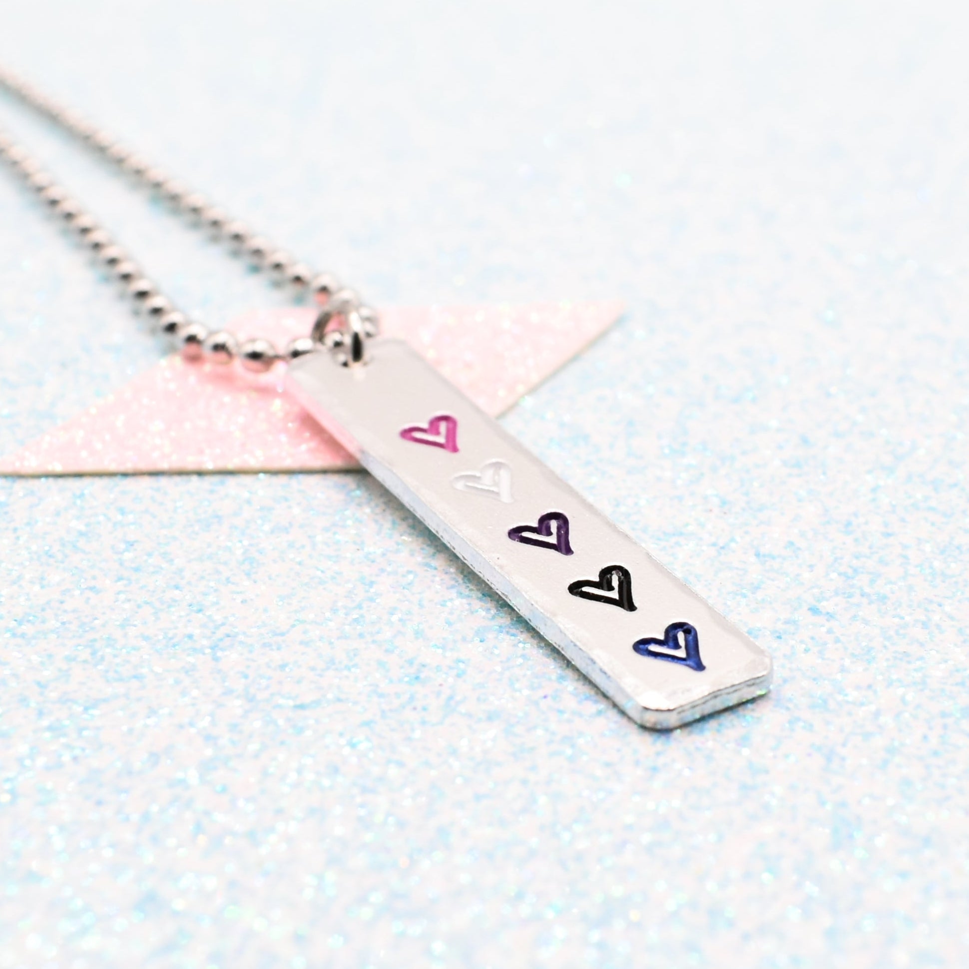 Necklace with a bar pendant on a ball chain featuring colourful hearts representing the gender fluid pride flag on a light blue background