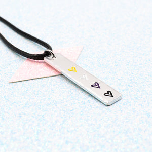 Necklace with a bar pendant on a faux leather cord featuring colourful hearts representing the non binary pride flag on a light blue background