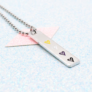 Necklace with a bar pendant on a ball chain featuring colourful hearts representing the non binary pride flag on a light blue background