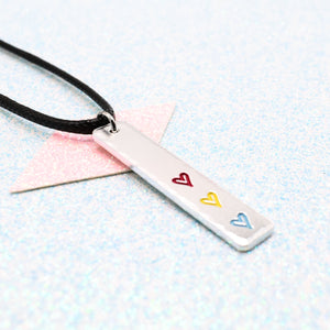 Necklace with a bar pendant on a faux leather cord featuring colourful hearts representing the pansexual pride flag on a light blue background