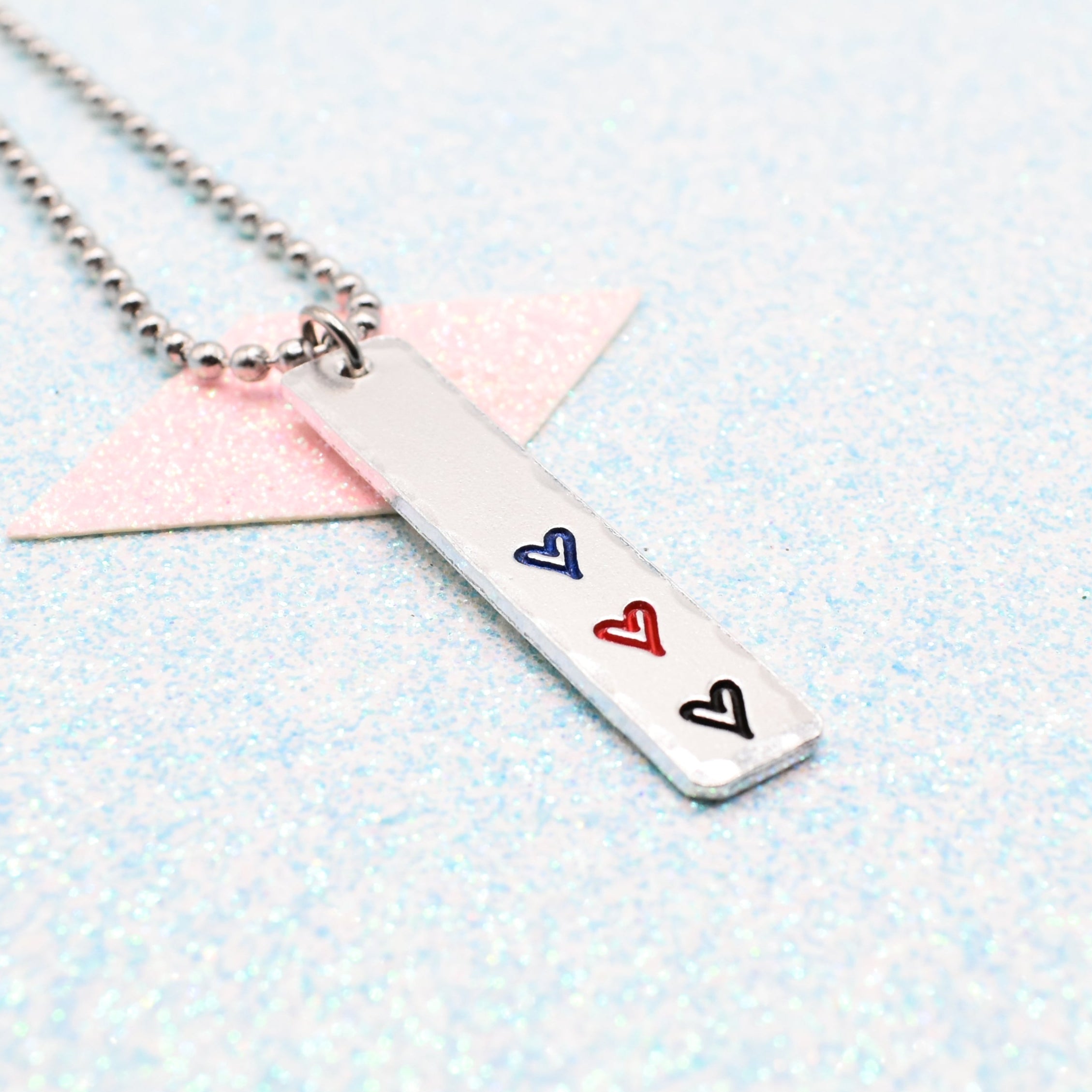 Necklace with a bar pendant on a ball chain featuring colourful hearts representing the polyamory pride flag on a light blue background