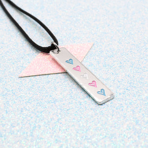 Necklace with a bar pendant on a faux leather cord featuring colourful hearts representing the transgender pride flag on a light blue background