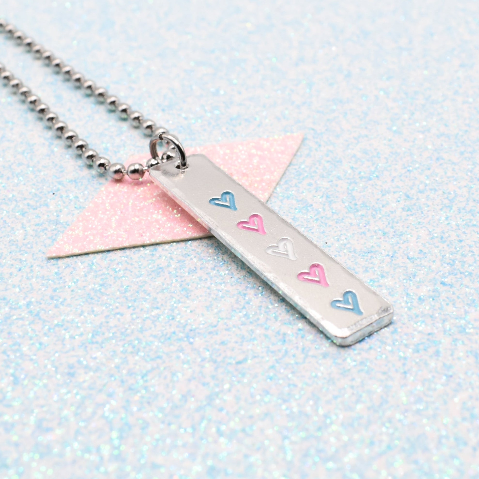 Necklace with a bar pendant on a ball chain featuring colourful hearts representing the transgender pride flag on a light blue background