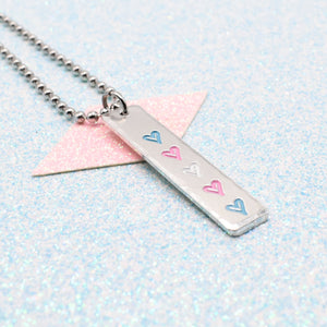 Necklace with a bar pendant on a ball chain featuring colourful hearts representing the transgender pride flag on a light blue background