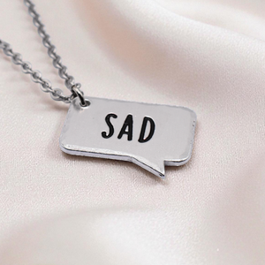 Silver necklace with a pendant that says 'SAD' on a light pink satin background
