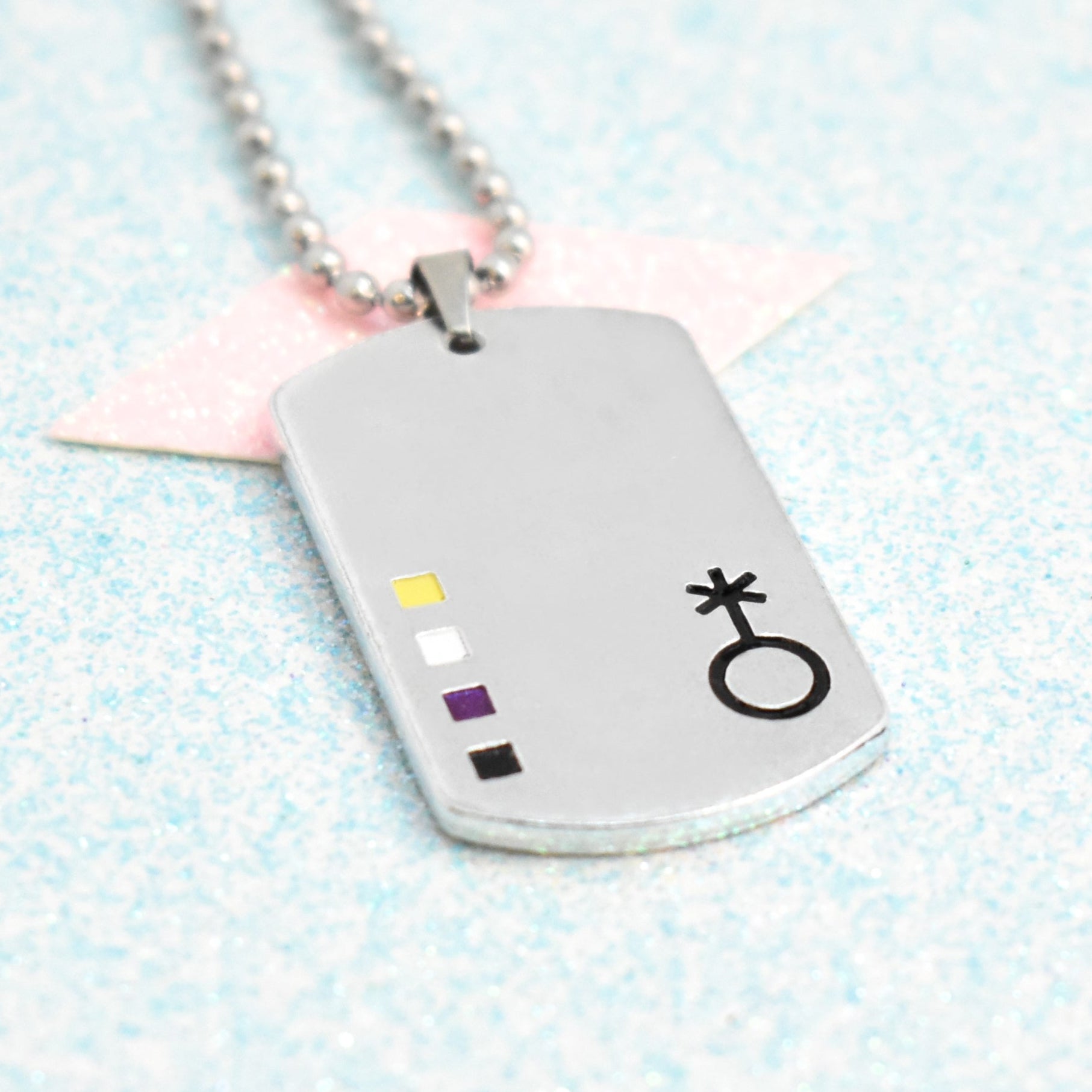 A silver-coloured necklace with a non-binary pride flag enamel pendant on a ball chain, displayed against a blue and pink glittery background.