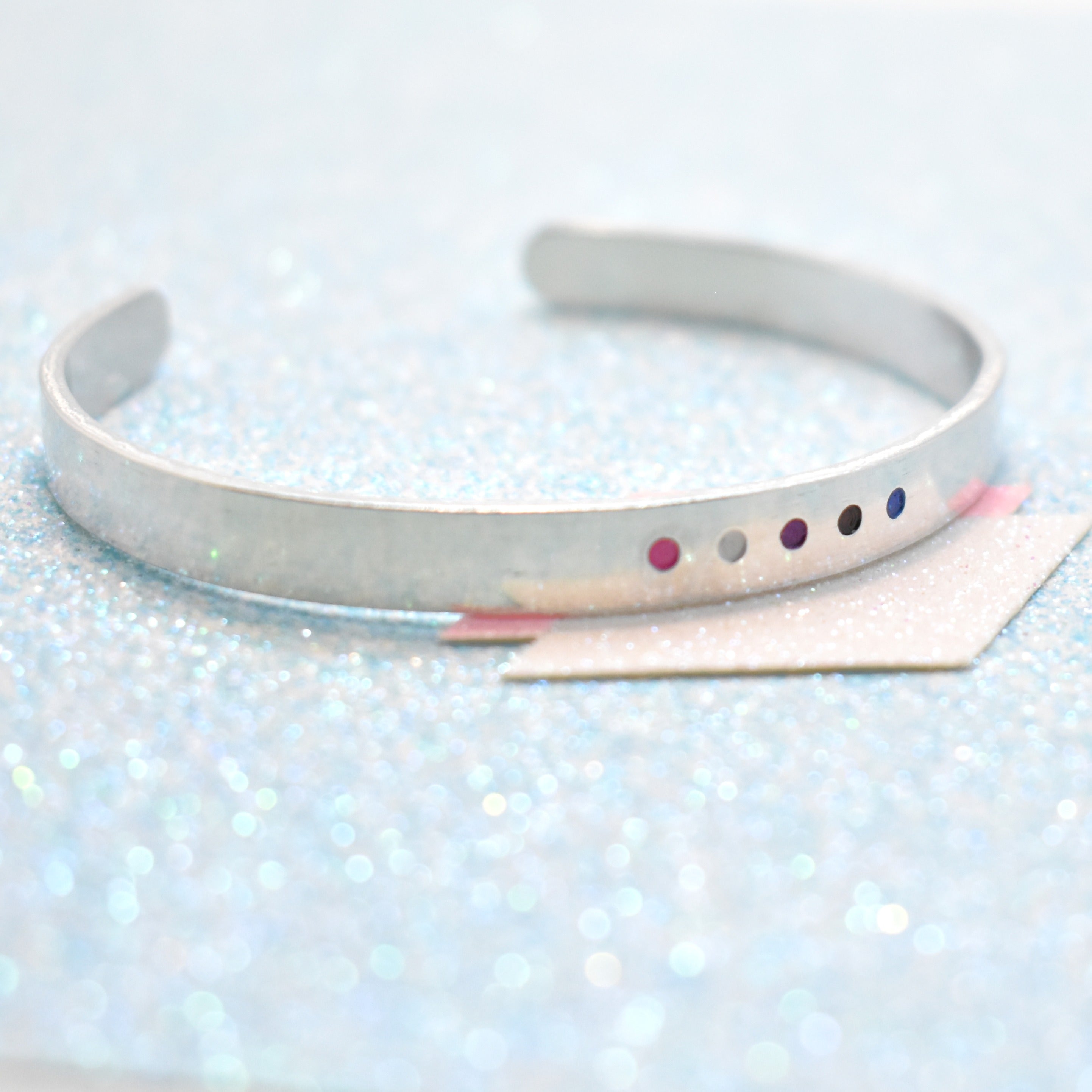 A bracelet with minimalist design, featuring colourful gender fluid accents, displayed on a glittery blue background.